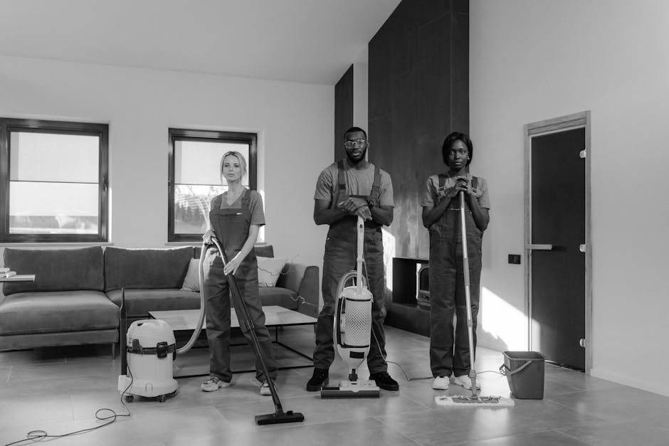 Three professional cleaners wearing grey t-shirts and red overalls stand indoors in a modern, well-lit room. The female cleaner on the left has blonde hair and holds a vacuum cleaner handle. The male 