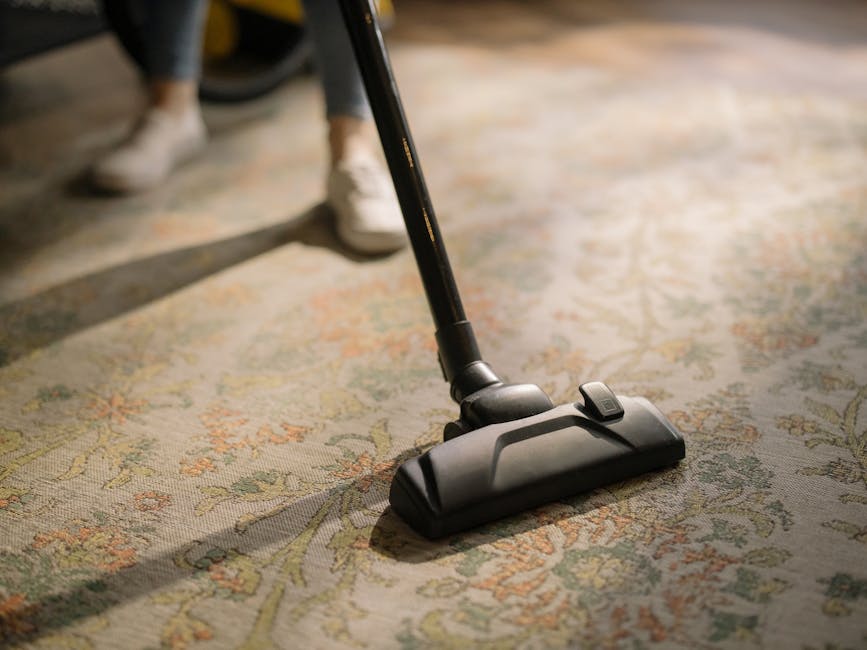 Close-up of a black vacuum cleaner head actively cleaning an intricately patterned, beige and pastel-colored carpet in a domestic setting, with a visible metal wand attached. The carpet surface appears clean and slightly textured, with natural lighting illuminating the area. In the background, part of a person's legs wearing dark pants and white shoes is visible, indicating active carpet maintenance. The scene conveys professional surface cleaning as provided by Carpet Cleaning E5, aligned with interior hygiene and deep cleaning practices in a residential space located on Chatsworth Road E5. The environment emphasizes routine carpet sanitisation and dust removal to maintain a fresh, well-maintained home interior.