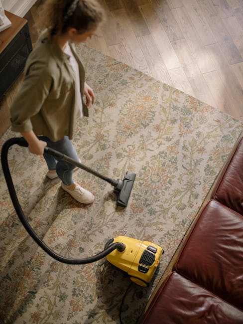 A woman using a yellow vacuum cleaner to perform deep surface cleaning on a patterned, light-colored carpet in a living room. She is dressed in casual clothing with a beige jacket, white sneakers, and has her hair pulled back. The room features a wooden floor adjacent to the carpet, with natural light illuminating the space. Alongside the carpet is a red leather sofa, and a radiator is visible at the top left corner. This scene showcases a professional cleaning process aimed at maintaining hygiene and freshness in a domestic setting, with visible cleaning equipment from Carpet Cleaning E5, a specialist in surface cleaning and sanitisation.