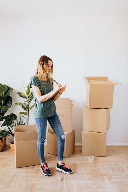 A young woman with shoulder-length blonde hair, dressed in a casual green t-shirt, ripped blue jeans, and black sneakers with pink accents, stands in a bright, minimally decorated room during daytime. She is holding a notepad and pen, inspecting or taking notes on her surroundings. Behind her are several empty cardboard boxes arranged in stacks, suggesting she is preparing for a move or cleaning process. To her left, there is a large indoor plant with broad green leaves in a woven basket, adding a touch of greenery to the space. The room has light-colored wooden flooring and a plain white wall, with natural light illuminating the scene. This image relates to professional domestic cleaning or end of tenancy cleaning services, as promoted by Carpet Cleaning E5, emphasizing thorough surface cleaning, decluttering, and hygienic preparation for property turnover.