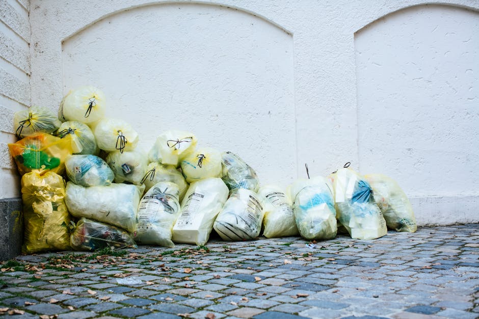 A young girl with dark curly hair tied back, wearing a yellow top, brown pleated skirt, white socks, and black shoes, is seen placing a crumpled plastic bag into a blue wheeled trash bin. The bin is s