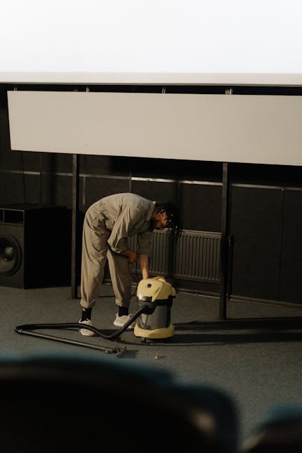 A person in a beige jumpsuit is performing surface cleaning in a commercial space, using a wet vacuum cleaner on a dark carpeted floor beneath a white acoustic ceiling panel. The room features black tiled walls, a radiator, and a large speaker, indicating a possible conference or event area. Bright, even lighting illuminates the scene, highlighting the cleanliness and maintenance activity. The image reflects professional deep cleaning procedures, consistent with services offered by Carpet Cleaning E5, aimed at maintaining hygiene and optimal surface condition in commercial settings.