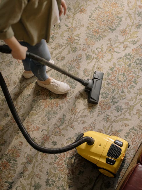A person dressed in a light-colored jumpsuit is performing surface cleaning or deep cleaning in an indoor environment, likely a conference or presentation room. They are bent over, operating a yellow