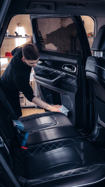 A professional cleaning technician from Carpet Cleaning E5 is inside a vehicle, cleaning the black leather seats with a cloth. The interior features dark leather upholstery with visible stitching, and the technician is focused on wiping the seat surface. The vehicle's back seats are clean and well-maintained, with a window in the background showing a blurred indoor environment. The scene highlights meticulous surface cleaning and attention to detail associated with domestic and vehicle upholstery cleaning services offered near London Fields Hackney, emphasizing hygiene and maintenance for comfortable driving and travel. Proper lighting illuminates the workspace, showcasing the cleanliness and condition of the car's interior.