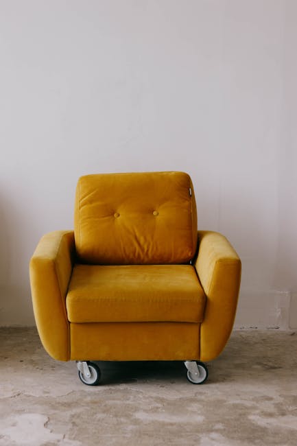 A professional cleaner wearing a dark jacket is using a handheld vacuum cleaner to deep clean a patterned yellow and white cushion placed on a dark grey fabric sofa within a residential room. The sofa's surface appears dust-free and well-maintained under bright, indoor lighting. The background features a plain white wall, highlighting the focus on surface cleaning and upholstery maintenance, with the image representing domestic upholstery sanitisation services offered by Carpet Cleaning E5 in London Fields Hackney.