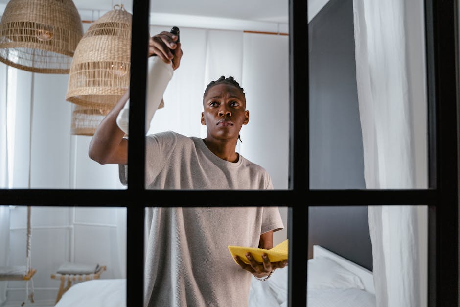 A woman with dark hair tied back is seen through a multi-pane window, wearing a light grey t-shirt and holding a yellow cloth in one hand while spraying cleaning solution onto a surface with the other